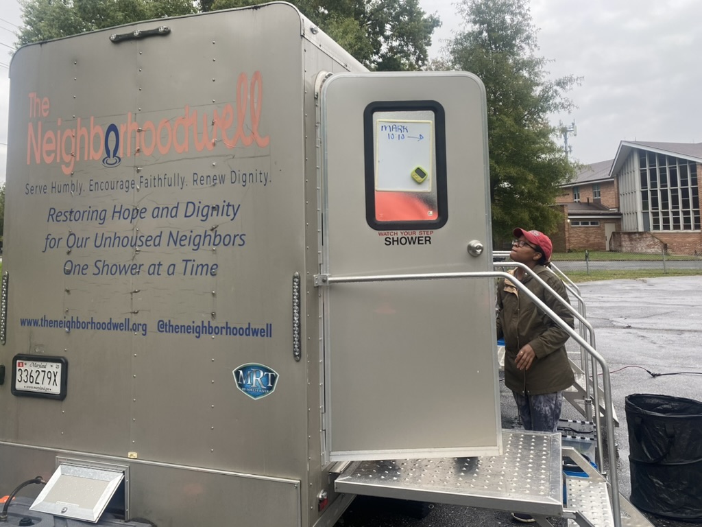 A Black woman stands at the entrance of a mobile silver shower.