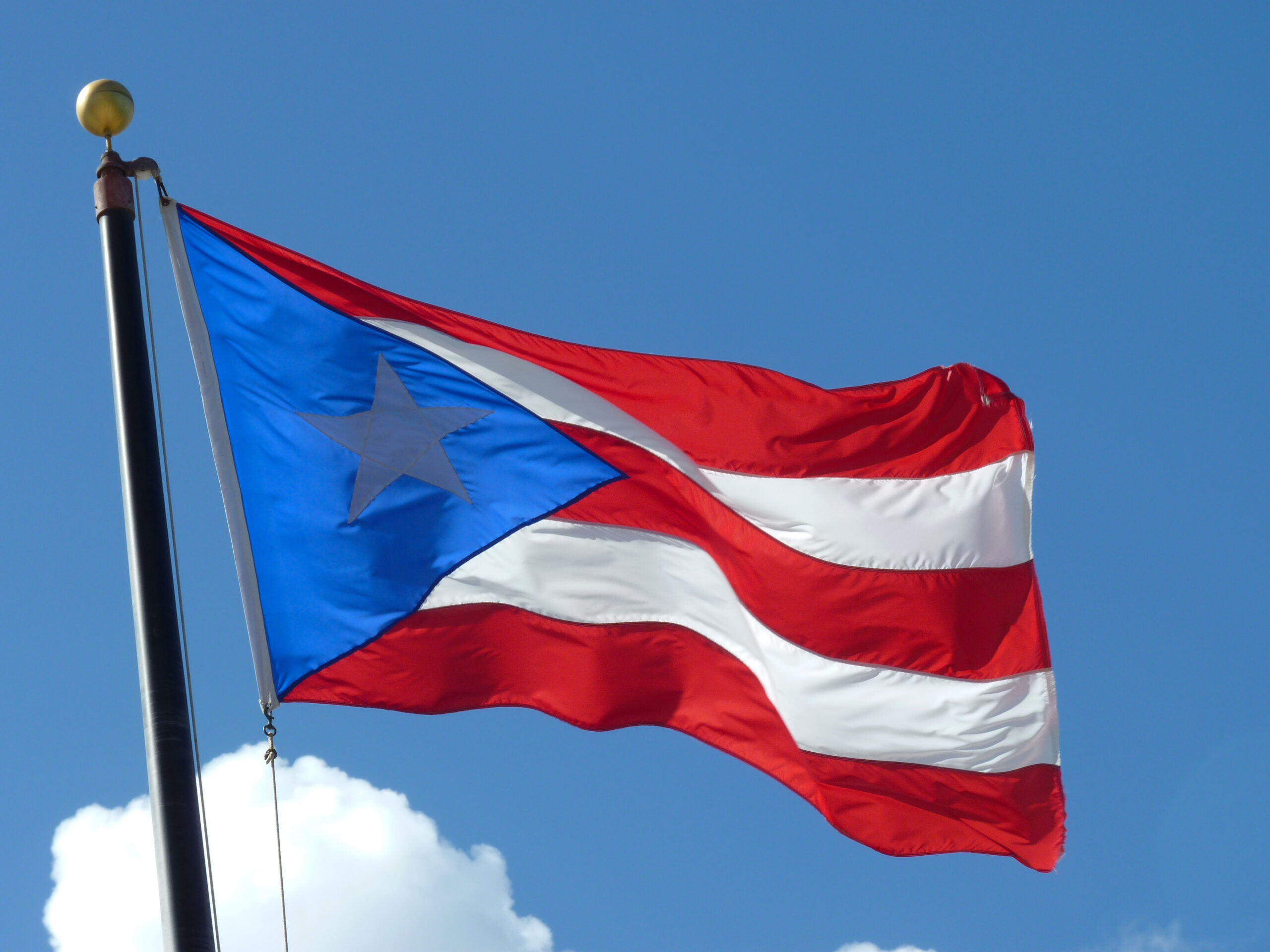 A red and white striped flag with a blue triangle flies in front of a blue sky.