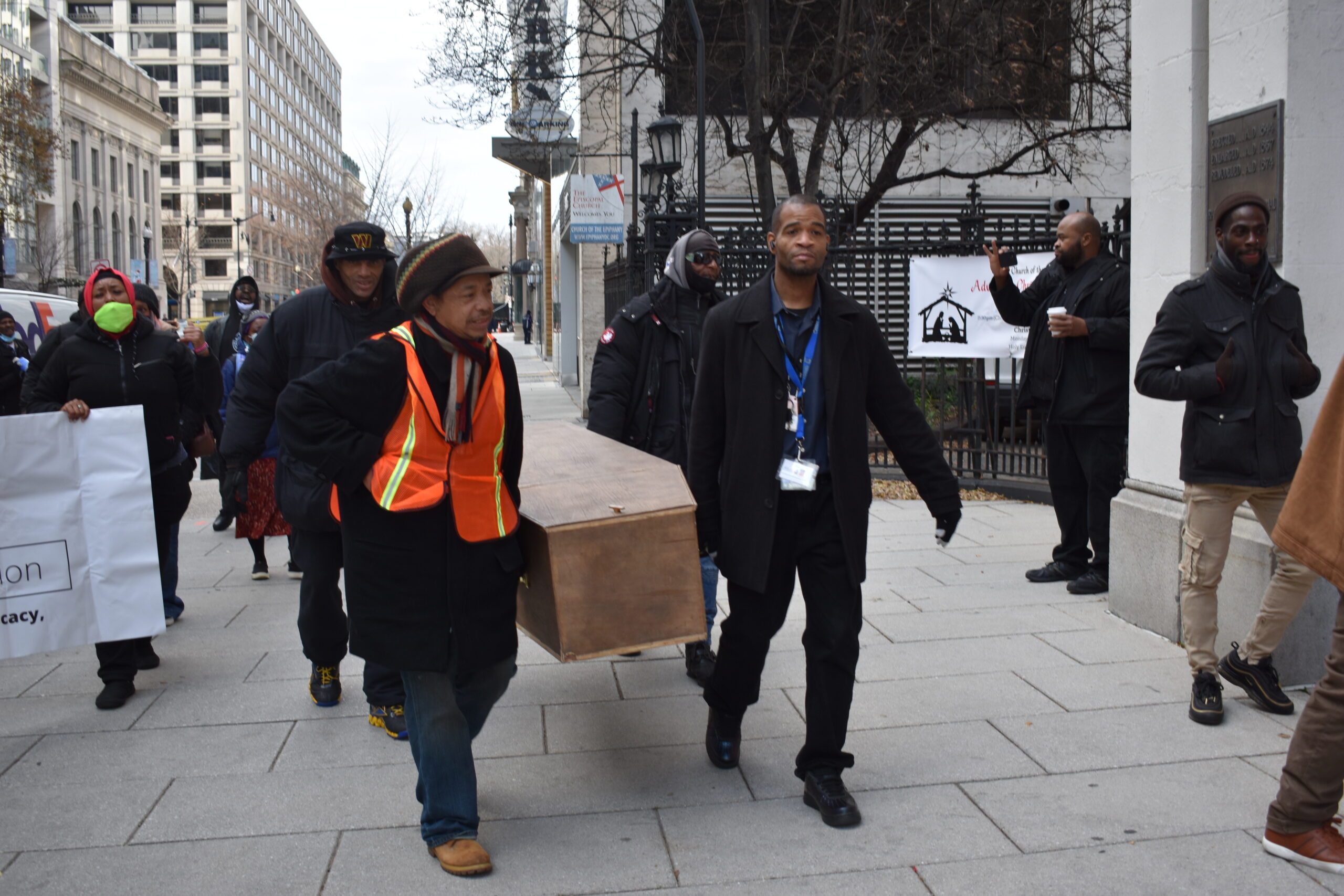 Two men carry a wooden coffin on the sidewalk, followed by a group of people.
