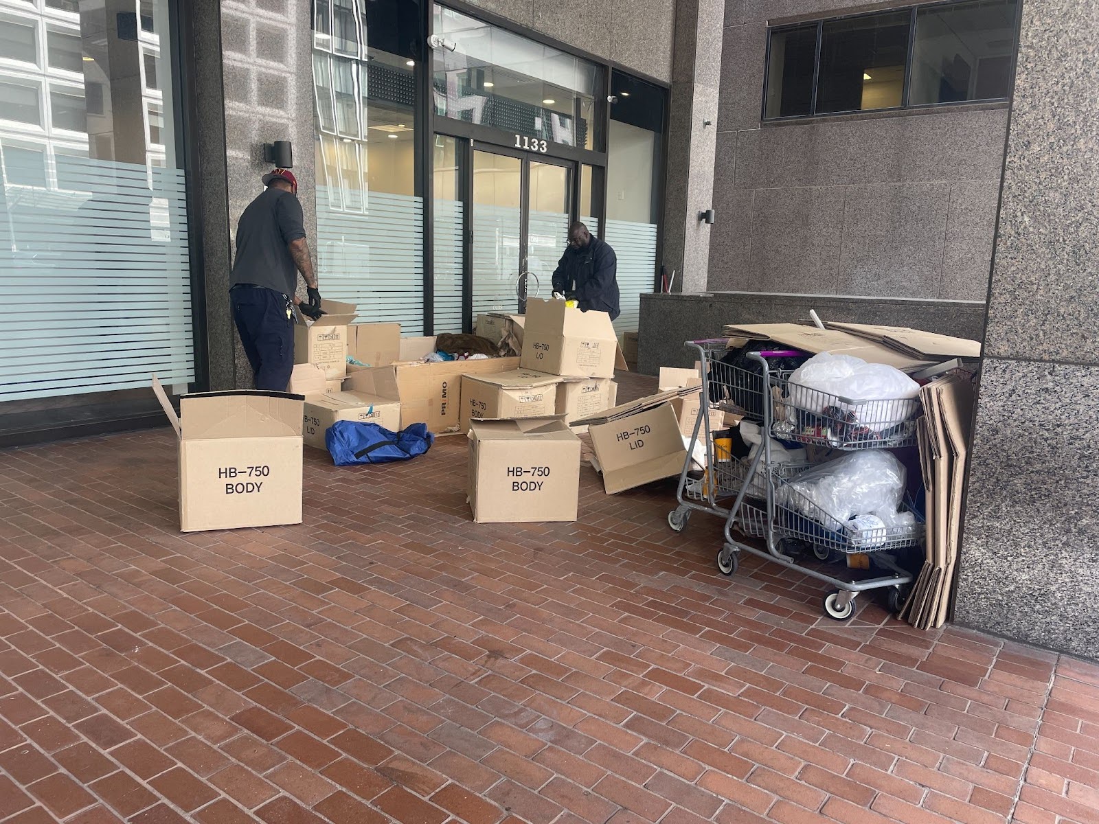 Two men go through cardboard boxes next to a grey building.