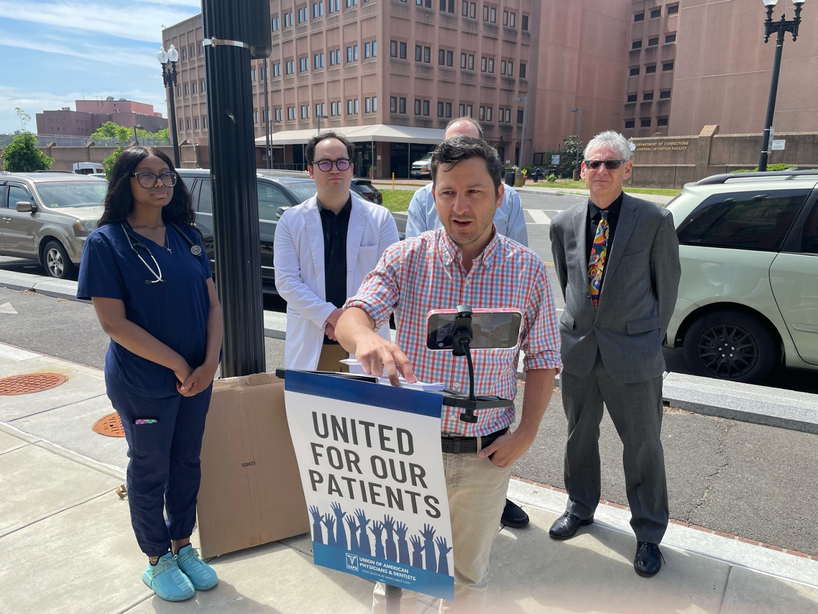 A group of fur people stand behind a speaker holding a sign that says "United for our patients."