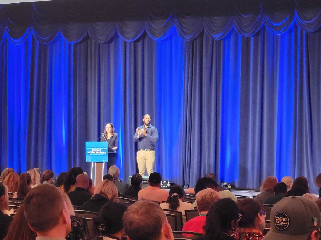 Women speaking at a podium in front of blue curtains.