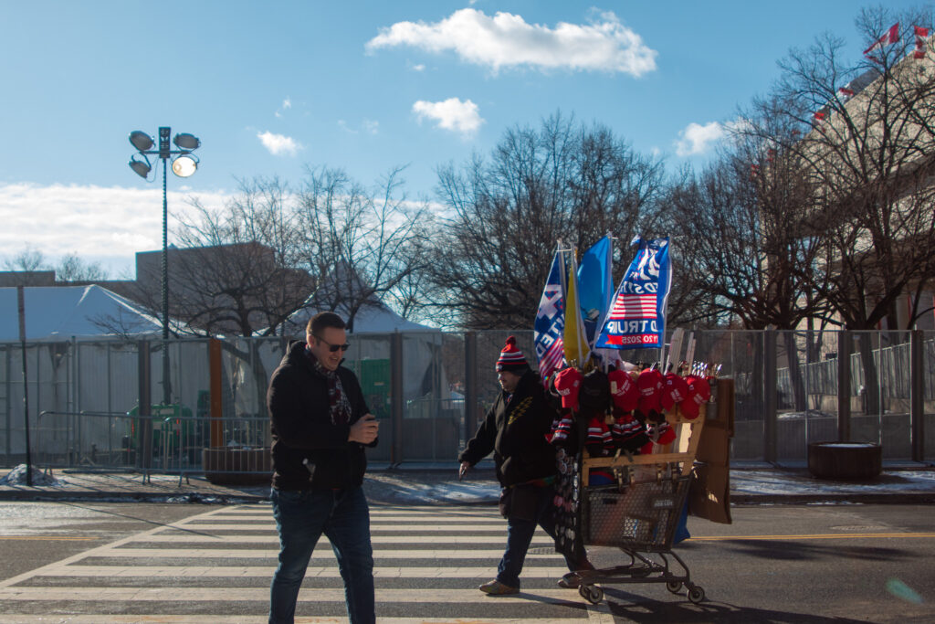 A man on his phone and a man with a cart of Donald Trump merchandise cross the street in front of a fence.