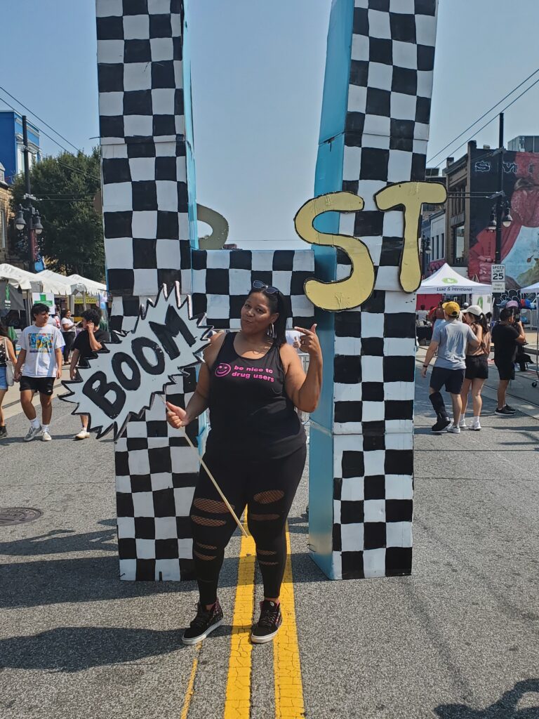 A woman poses in all black holding a sign that says "Boom."
