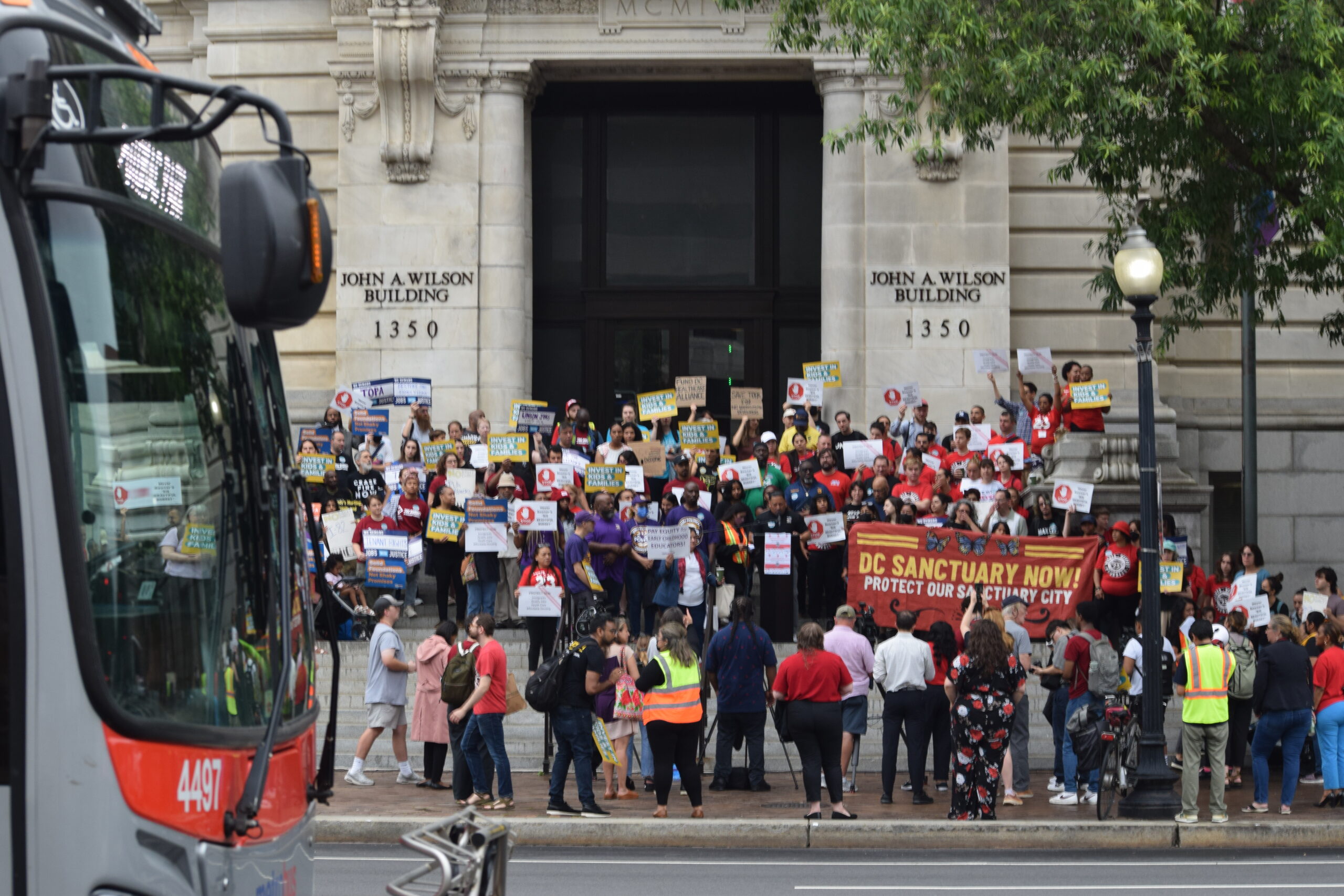 A group of people wearing colorful shirts and holding signs about the budget stand in front of D.C.'s city building.