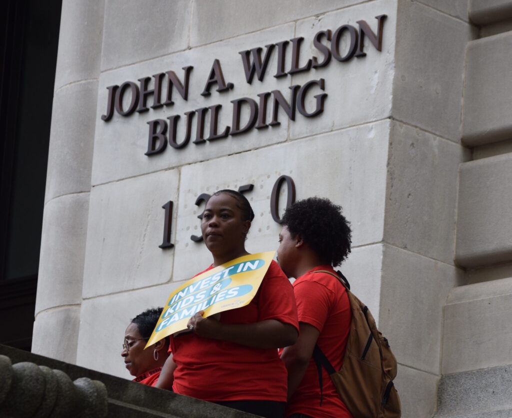 Two women holding signs stand in front of the D.C. Council building.