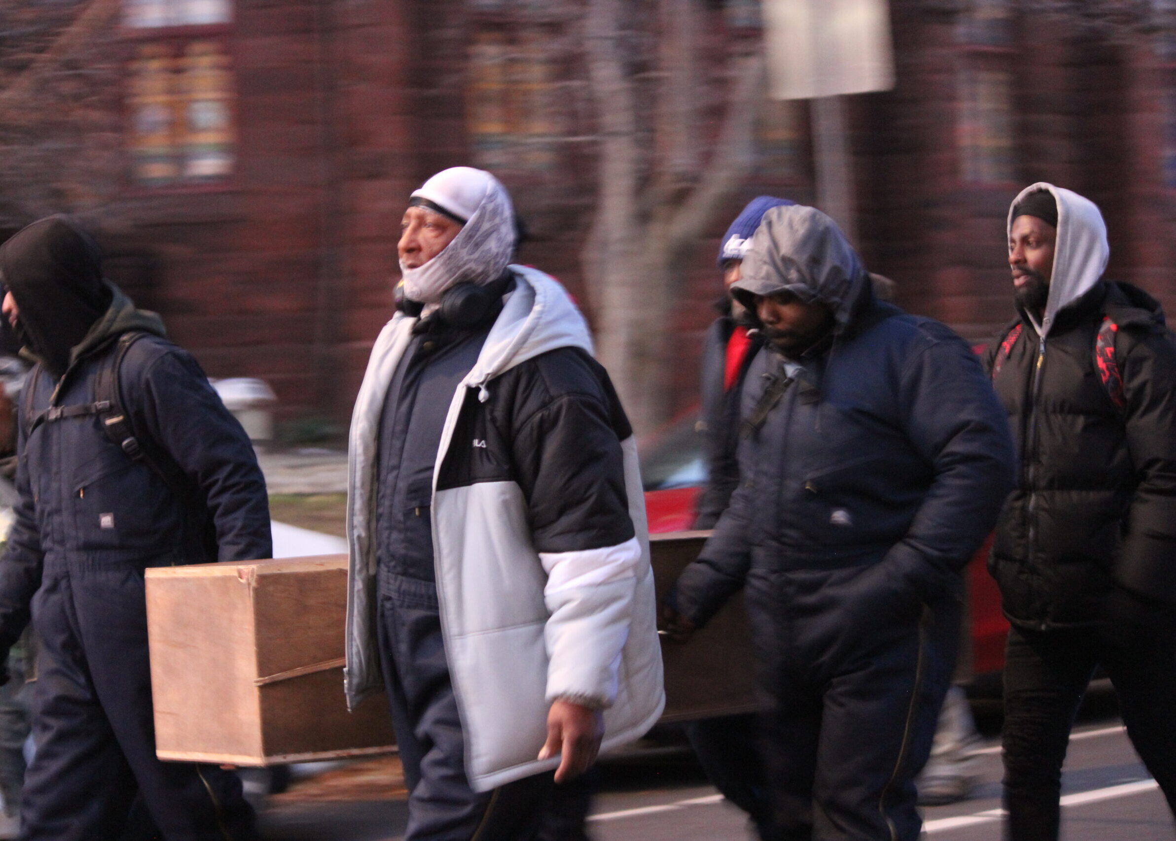 Five men in coats carry a wooden coffin outside